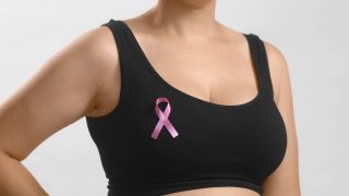 jovem de blusa preta em um fundo branco exibindo um laço cor-de-rosa no peito em apoio ao Outubro Rosa e às pessoas que enfrentam o câncer de mama